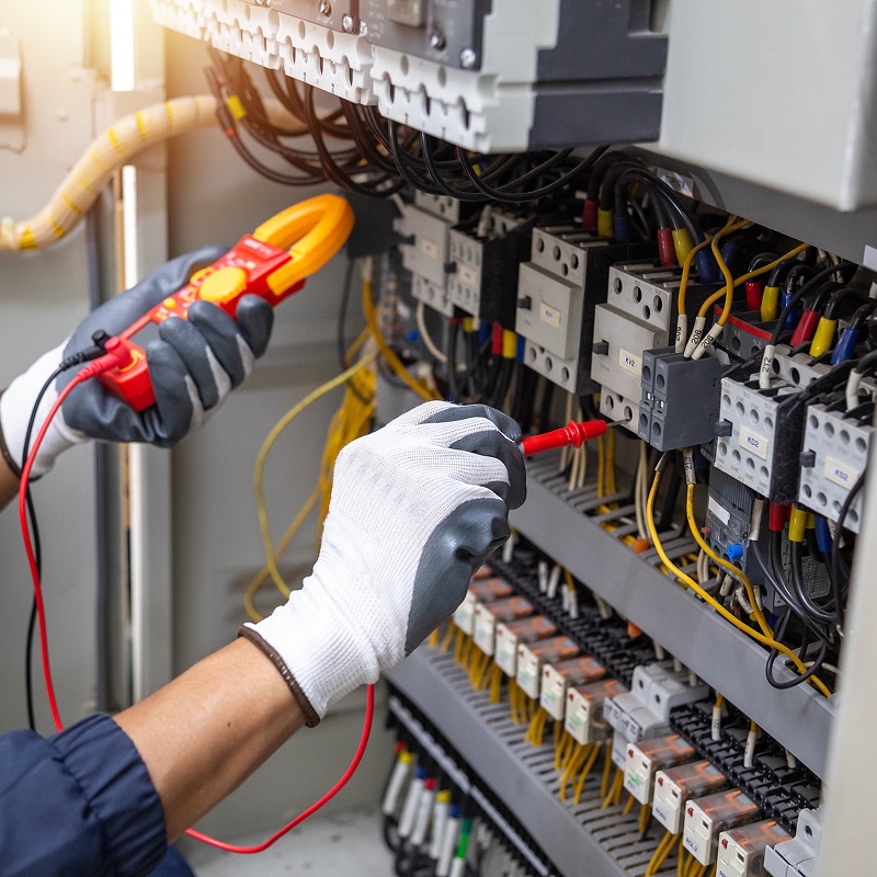 Électricien en gants testant un tableau électrique avec un multimètre à pince, mesurant les composants. Sécurité.