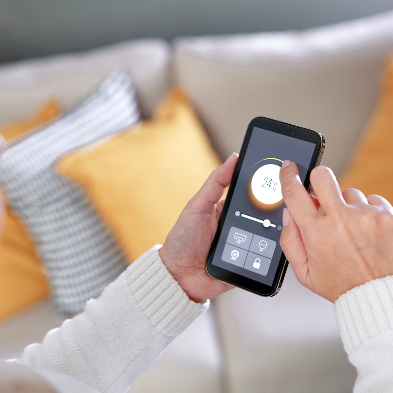 Mains ajustant 24°C sur un smartphone avec appli maison connectée : contrôle chauffage, lumière, sécurité, serrure. Canapé en arrière-plan.